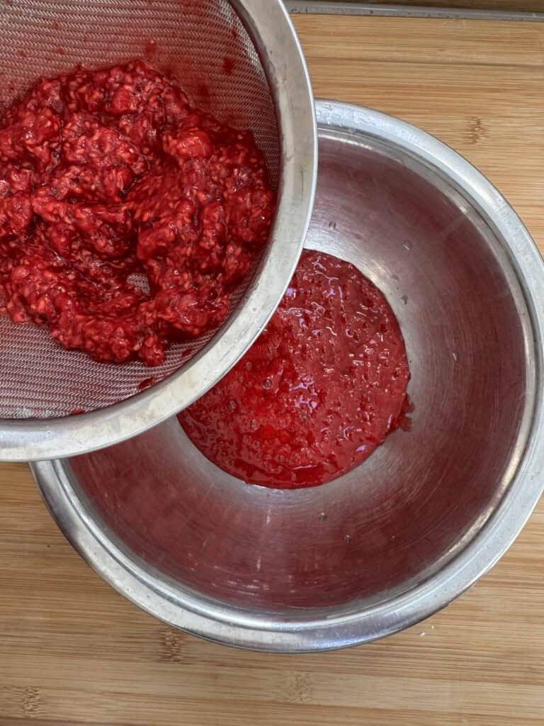A metal strainer holds mashed red raspberries over a bowl, with juice draining into another metal bowl below, all set on a wooden surface.