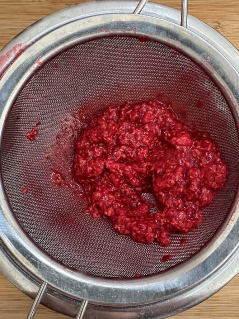 Bright red mashed raspberries rest in a fine mesh strainer over a metal bowl, with juice seeping through the strainer. The setup is on a wooden surface, suggesting food preparation.