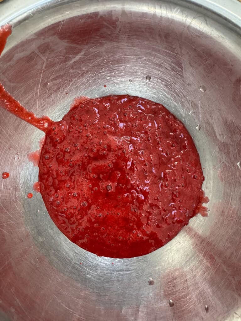 A stainless steel bowl containing a thick, red, slightly chunky liquid, resembling blended or pureed fruit, with some splatters on the edge of the bowl.