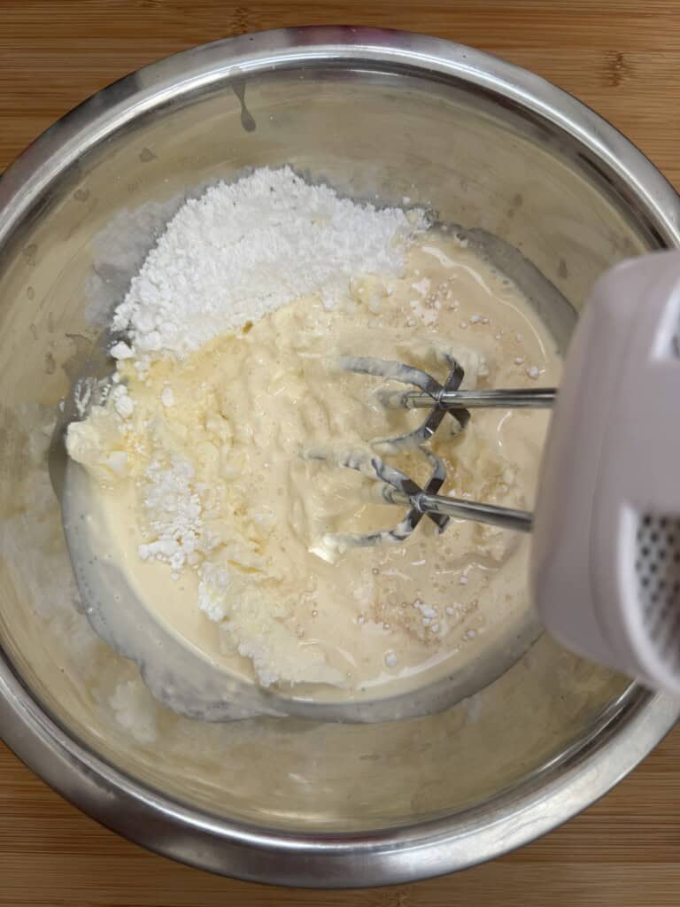 A close-up of a metal mixing bowl containing cream, powdered sugar, and other ingredients being blended with an electric hand mixer on a wooden surface.