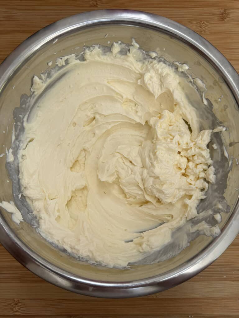 A large stainless steel bowl filled with smooth, creamy, pale yellow frosting or whipped cream, with swirls visible on the surface, sits on a wooden countertop.