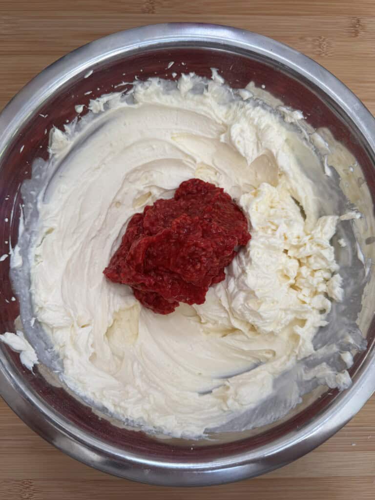A metal bowl filled with creamy white mixture, likely cream cheese, with a dollop of bright red raspberry puree on top, sitting on a wooden surface.