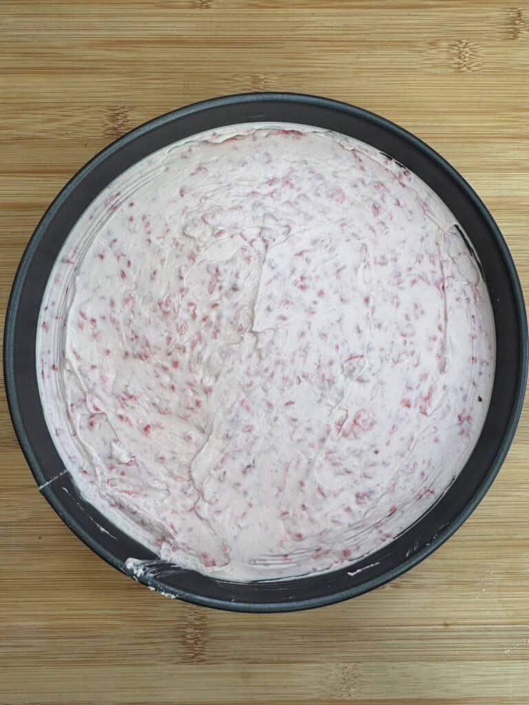 A round baking pan filled with an even layer of pale pink, raspberry-speckled cheesecake mixture sits on a light wooden surface.