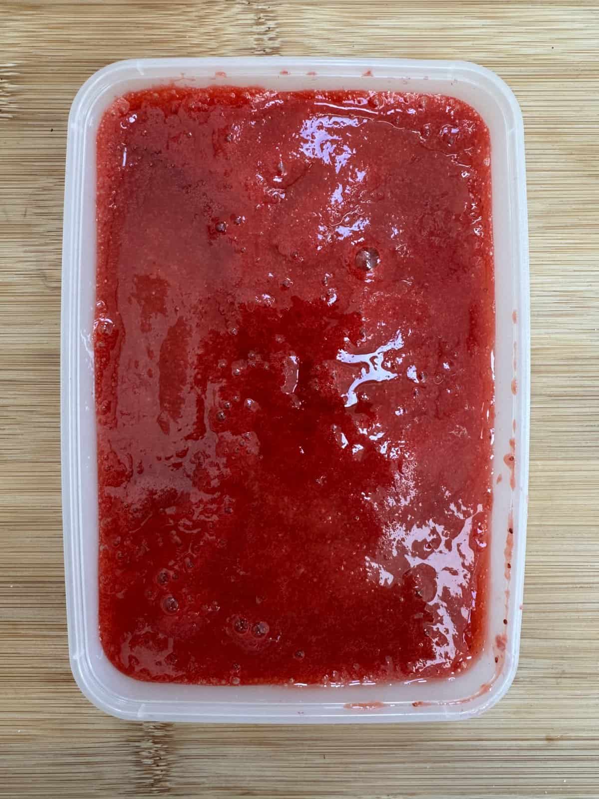 A rectangular plastic container filled with bright red strawberry puree sits on a light wooden surface.
