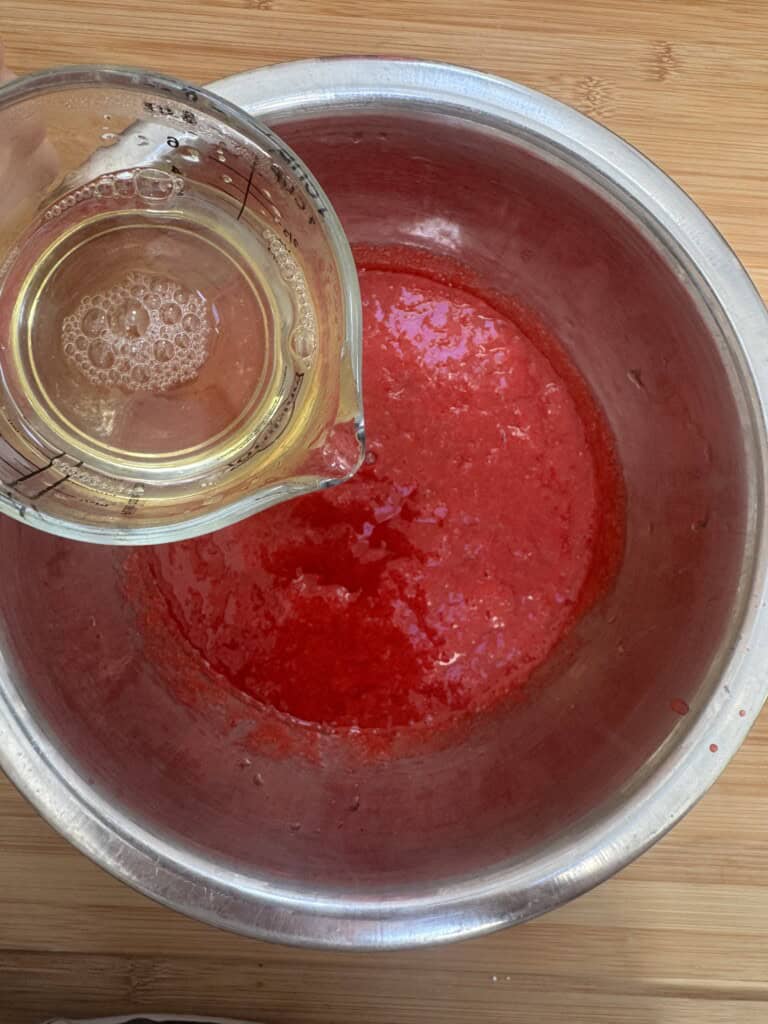 A glass measuring cup pours a clear liquid into a metal bowl containing a bright red mixture, set on a wooden surface.