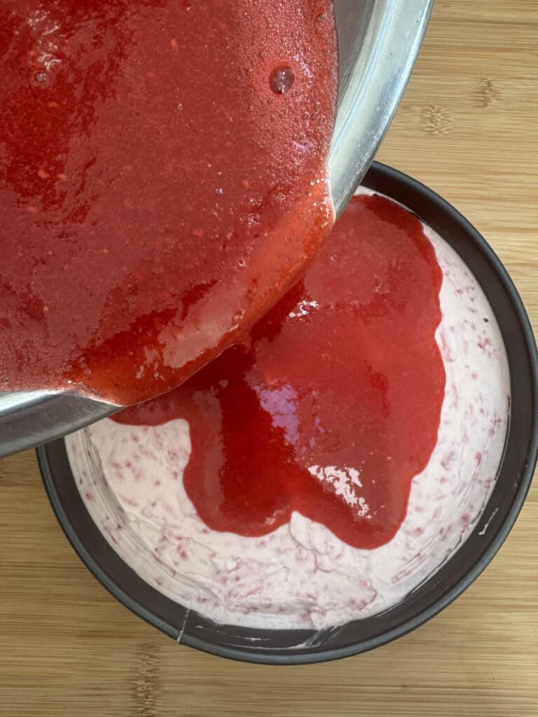 A close-up of red strawberry sauce being poured from a metal bowl over a round cake with a creamy, pale pink filling in a cake pan, set on a wooden surface.