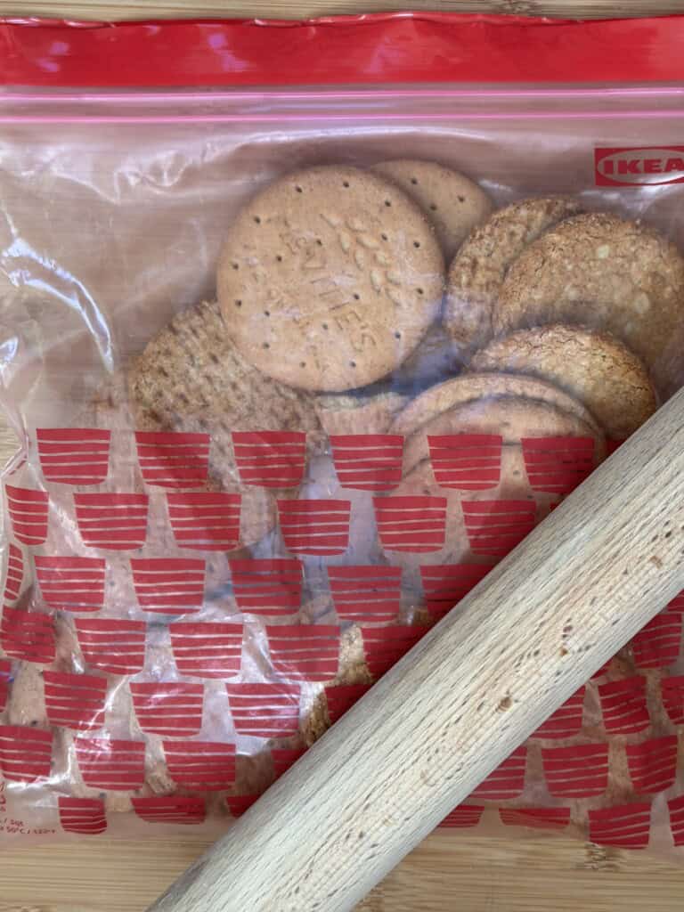 A clear plastic IKEA bag with red patterns holds round biscuits. In the foreground, a wooden rolling pin lies diagonally across the bag on a wooden surface.