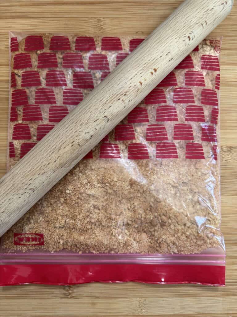 A rolling pin rests on a sealed plastic bag filled with crushed graham crackers, set on a wooden surface.