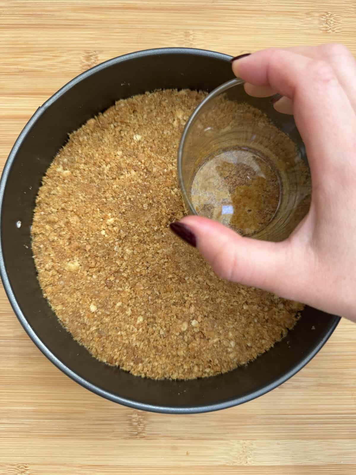 A hand presses a glass cup into a round pan filled with crushed graham cracker crust on a wooden surface, preparing a dessert base.