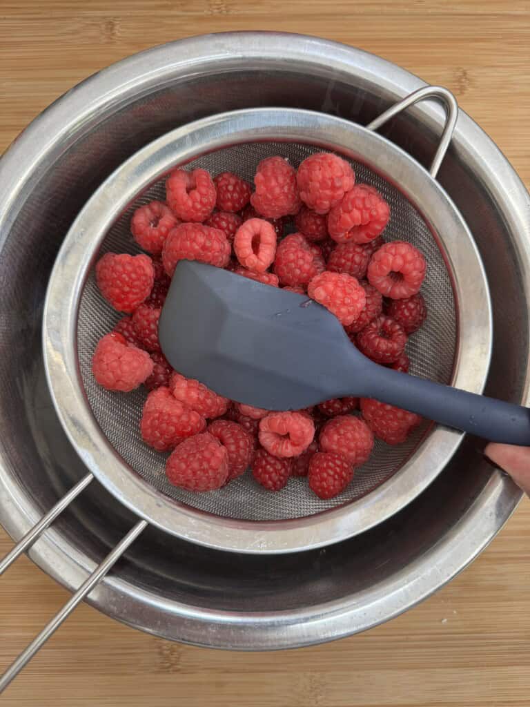A gray spatula presses fresh raspberries through a fine mesh strainer placed over a metal bowl, all set on a wooden surface.
