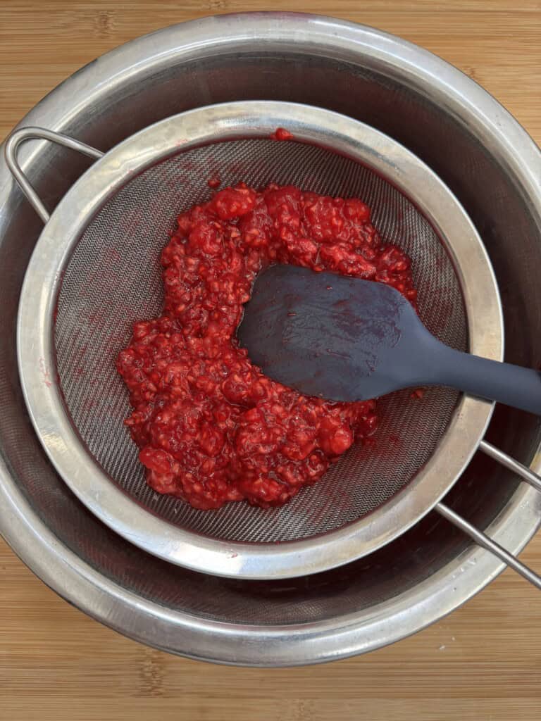 A black spatula presses mashed raspberries through a metal strainer set over a large steel bowl on a wooden surface.