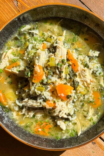 A bowl of Scottish chicken and rice soup with shredded chicken, carrots, celery, herbs, and rice in a broth, served on a wooden table in natural sunlight.