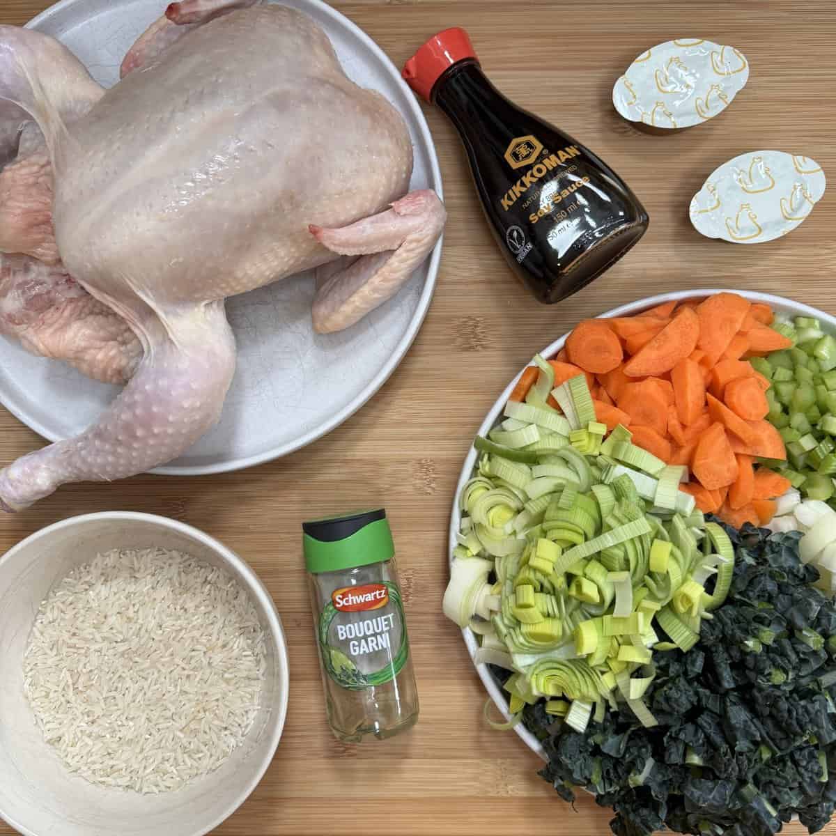 Ingredients for Scottish Chicken and Rice Soup. A whole raw chicken on a plate, a bowl of uncooked rice, a bottle of soy sauce, two bouillon tablets, a jar of bouquet garni seasoning, and a plate with chopped carrots, celery, leeks, and leafy greens on a wooden surface.