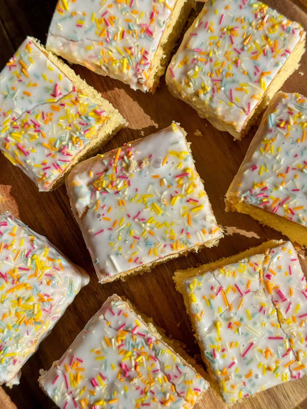 Several square slices of cake are topped with white icing and colorful sprinkles, arranged on a wooden surface in bright natural light.