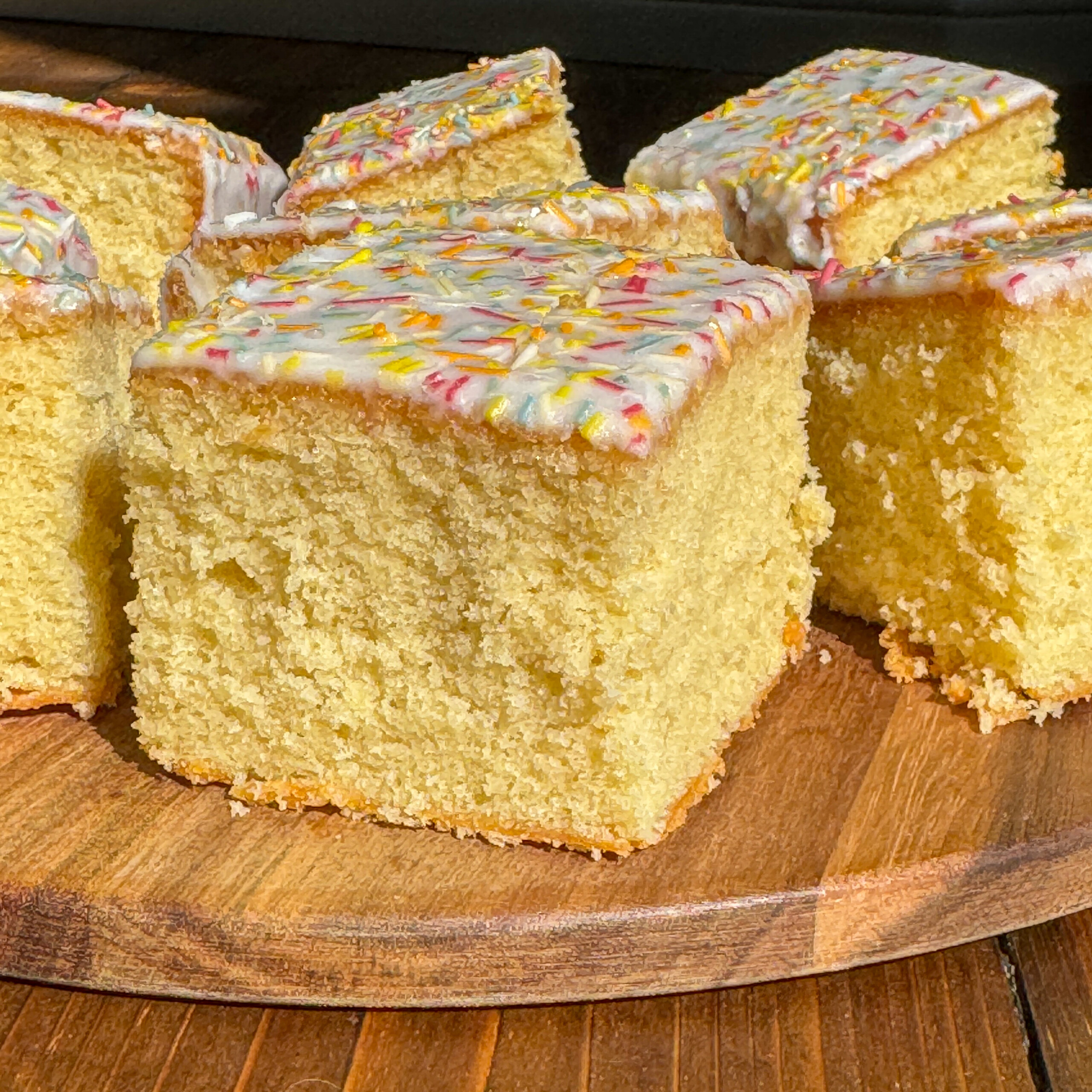 Several pieces of primary school cake with white icing and sprinkles are arranged on a wooden platter in bright natural light.