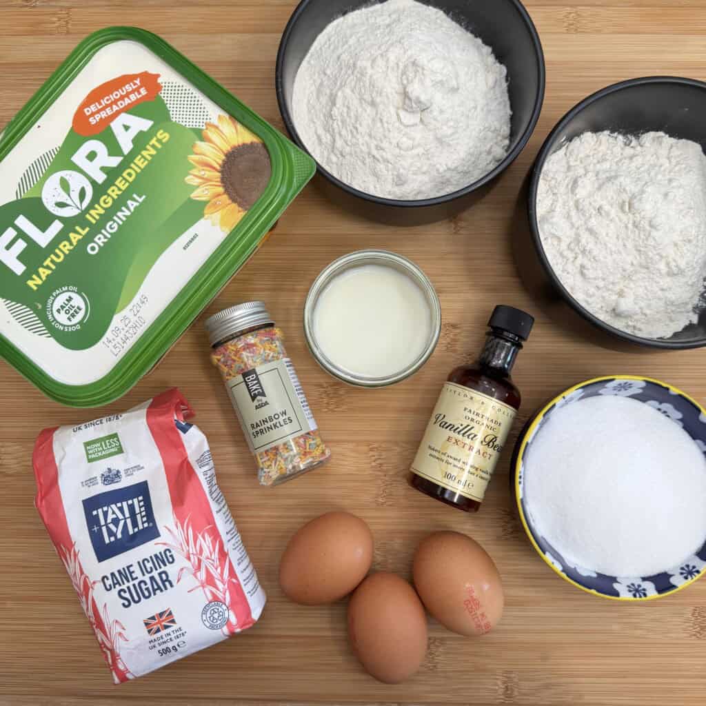 Baking ingredients on a wooden surface: margarine tub, two bowls of flour, a bowl of sugar, vanilla extract bottle, milk, rainbow sprinkles, icing sugar pack, and three eggs.