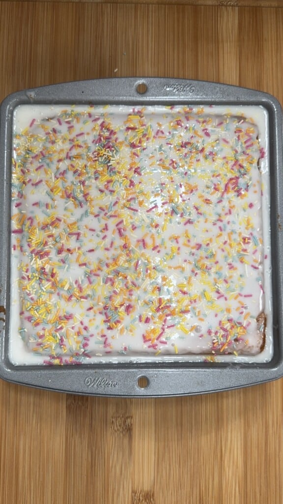 A square cake in a gray baking pan, topped with white icing and colorful sprinkles, sits on a wooden surface.
