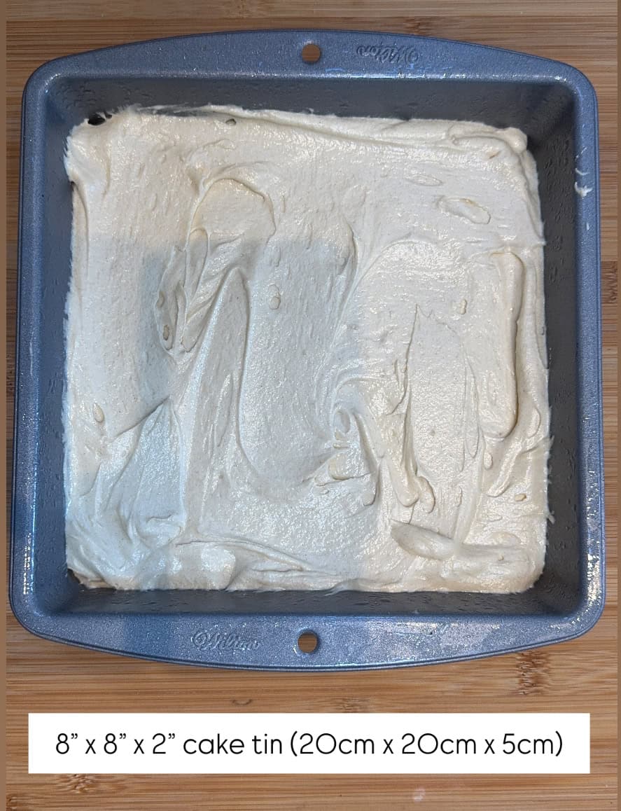 A square metal cake tin, 8x8 inches, filled with unbaked cake batter, shown from above. A label at the bottom reads 8” x 8” x 2” cake tin (20cm x 20cm x 5cm).