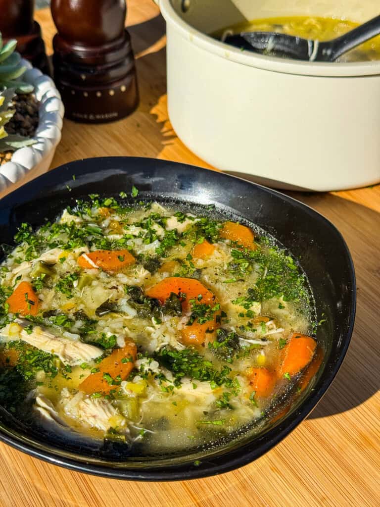 A black bowl of chicken and rice soup with carrots, herbs, and shredded chicken sits on a wooden table, with a white pot of soup and pepper mills in the background. Sunlight highlights the fresh ingredients.