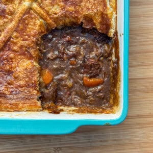 A rectangular blue baking dish with a golden-brown pastry-topped Scottish steak pie. A portion has been removed, revealing a rich, dark filling with chunks of meat and carrots on a wooden surface.