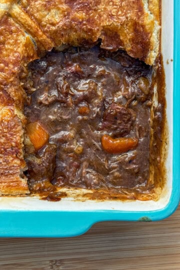 A rectangular blue baking dish with a golden-brown pastry-topped slow cooker steak pie. A portion has been removed, revealing a rich, dark filling with chunks of meat and carrots on a wooden surface.
