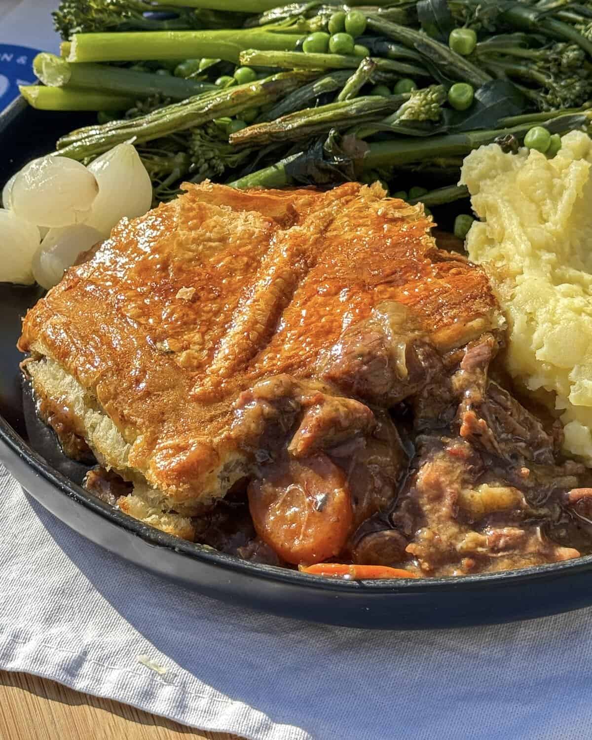 A close-up of slow cooker steak pie with flaky crust, served with mashed potatoes, glazed onions, and broccolini on a black plate. The pie is cut open, revealing a rich filling with carrots and gravy inside.