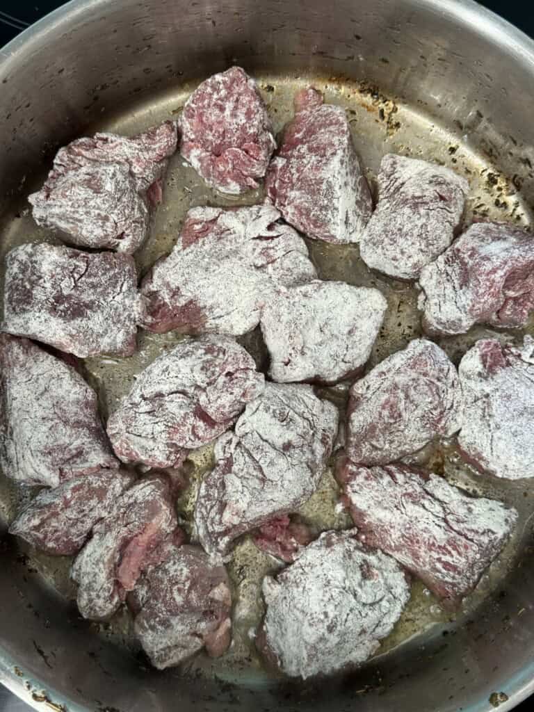Chunks of raw beef coated in flour are arranged in a single layer in a stainless steel pan, ready to be browned or cooked. Some browned spots are visible on the pan’s surface.