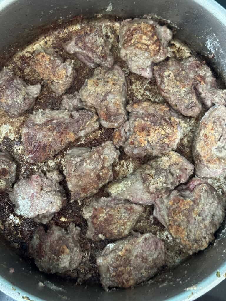 Several browned pieces of meat are cooking in a large pan, with visible seasoning and a crispy, seared surface. Some areas of the pan have rendered fat and browned residue.