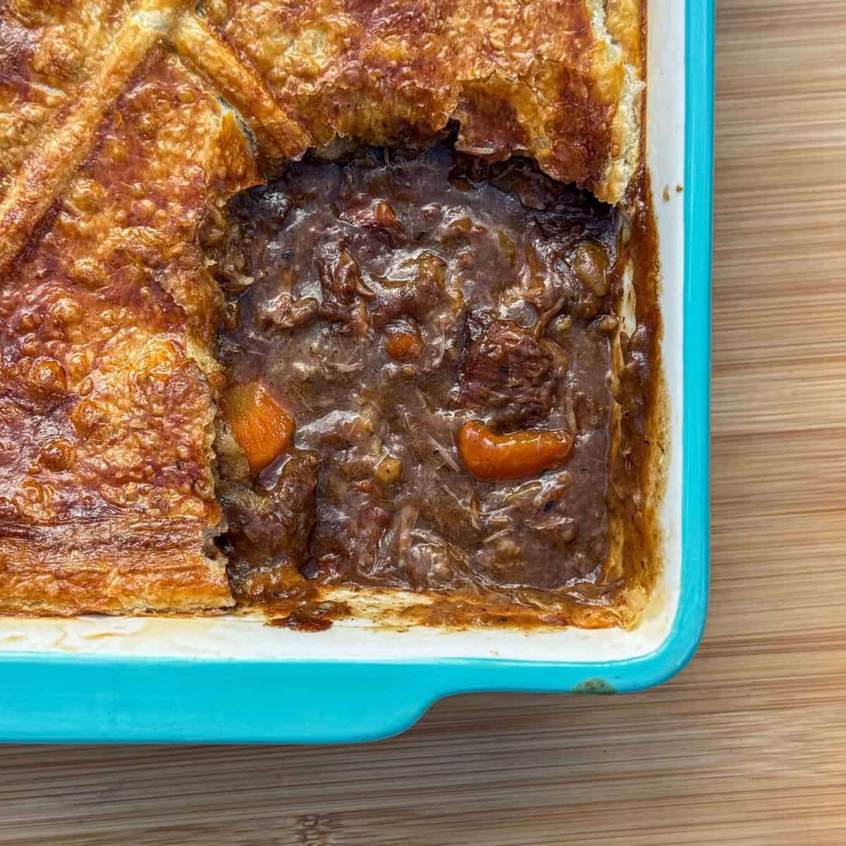 A rectangular blue baking dish with a golden-brown pastry-topped Scottish steak pie. A portion has been removed, revealing a rich, dark filling with chunks of meat and carrots on a wooden surface.