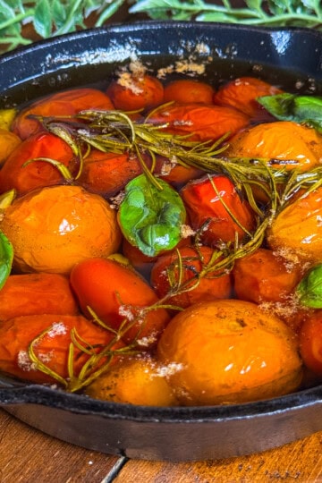 A black cast iron skillet filled with confit tomatoes, fresh basil leaves, and sprigs of rosemary in olive oil, sitting on a wooden surface with green foliage in the background.