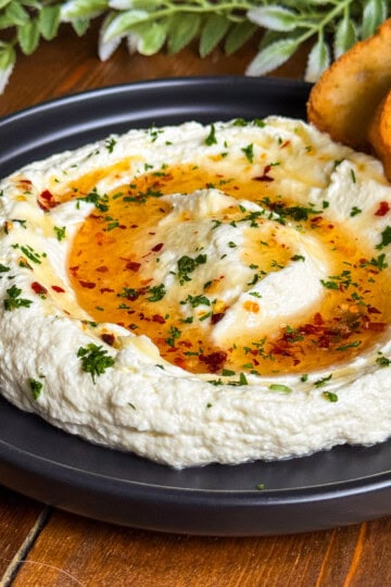 A black plate of creamy whipped feta garnished with hot honey chopped herbs, and red pepper flakes, served with two toasted bread slices. Green leafy decor is in the background.