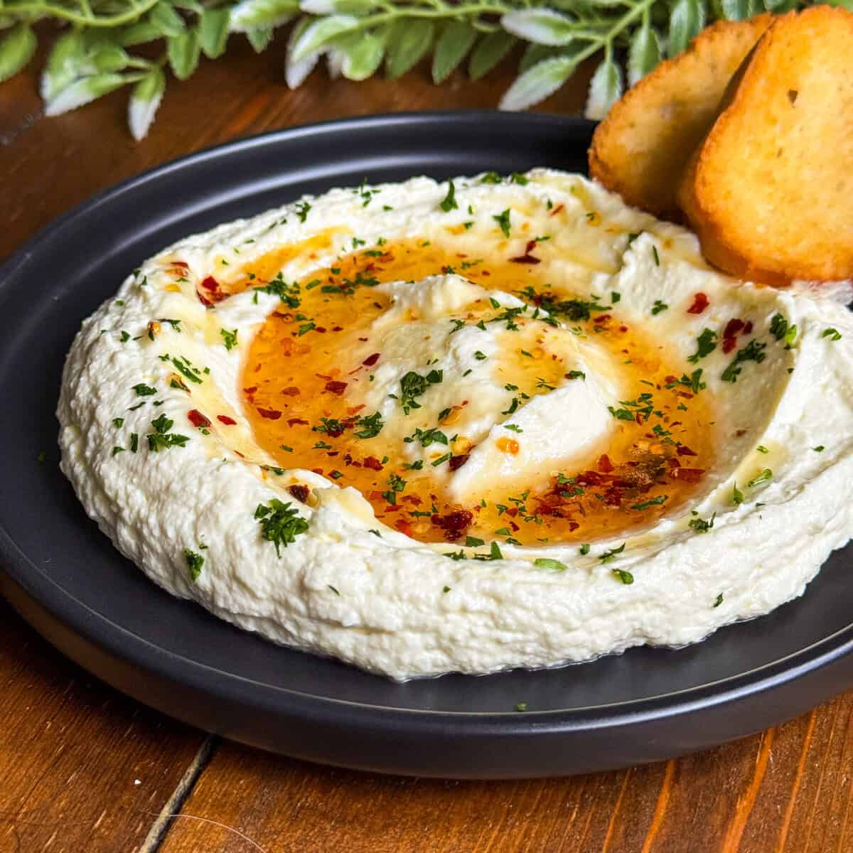 A black plate of creamy whipped feta garnished with hot honey chopped herbs, and red pepper flakes, served with two toasted bread slices. Green leafy decor is in the background.