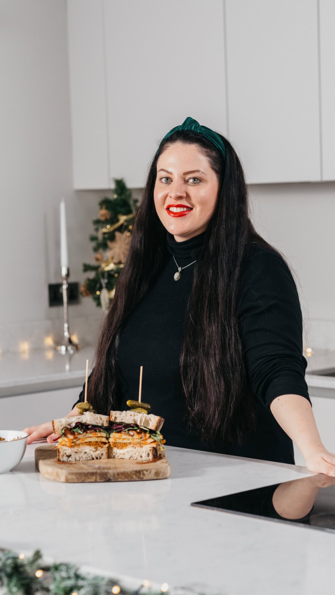 A woman with long dark hair and red lipstick stands in a modern kitchen, smiling beside a cutting board with stacked sandwiches. There is a festive plant and a candle in the background.