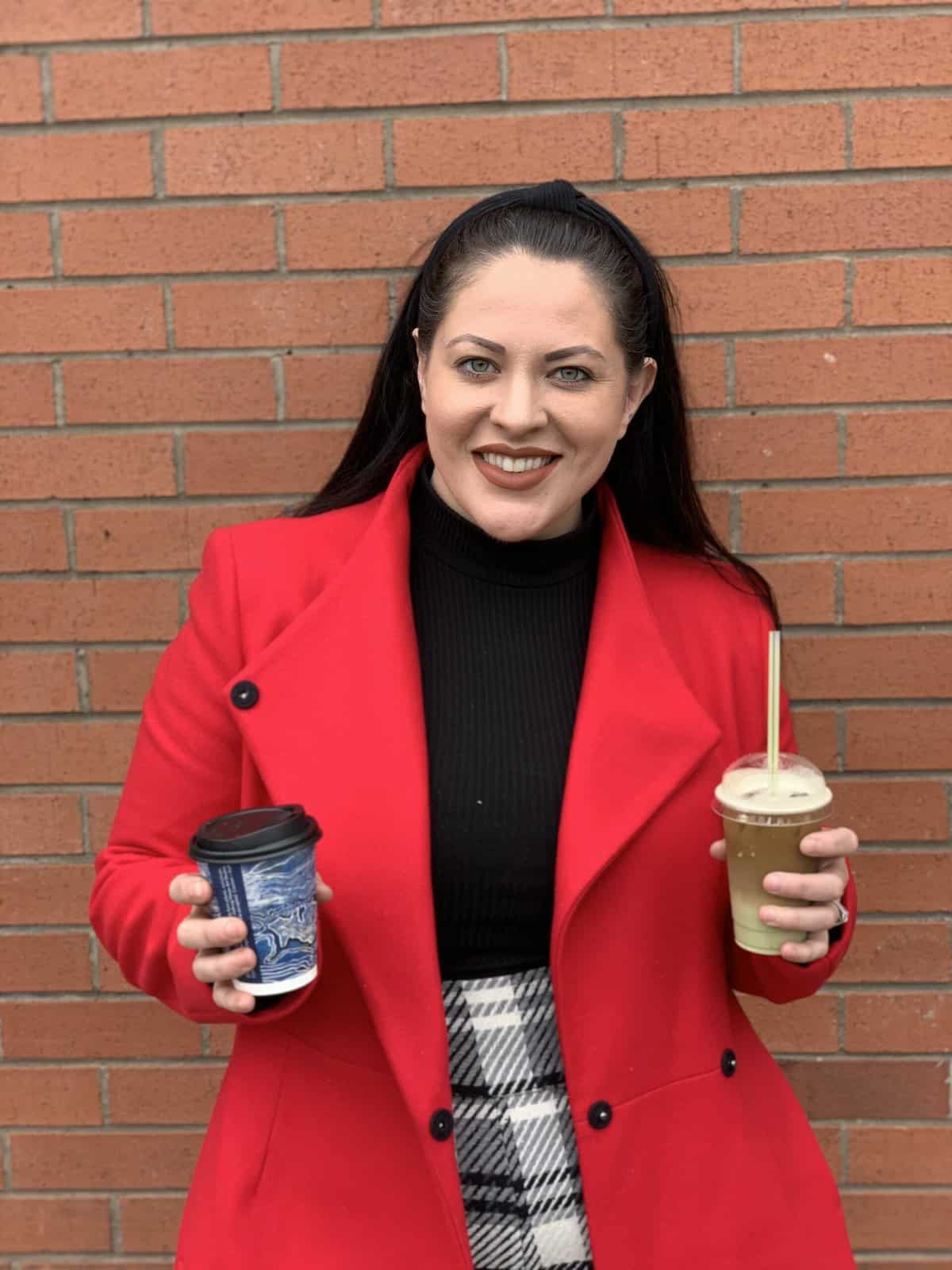 A woman with long dark hair in a high ponytail wears a bright red coat, black turtleneck, and plaid skirt. She smiles while holding a hot drink in one hand and an iced drink in the other, standing in front of a brick wall.