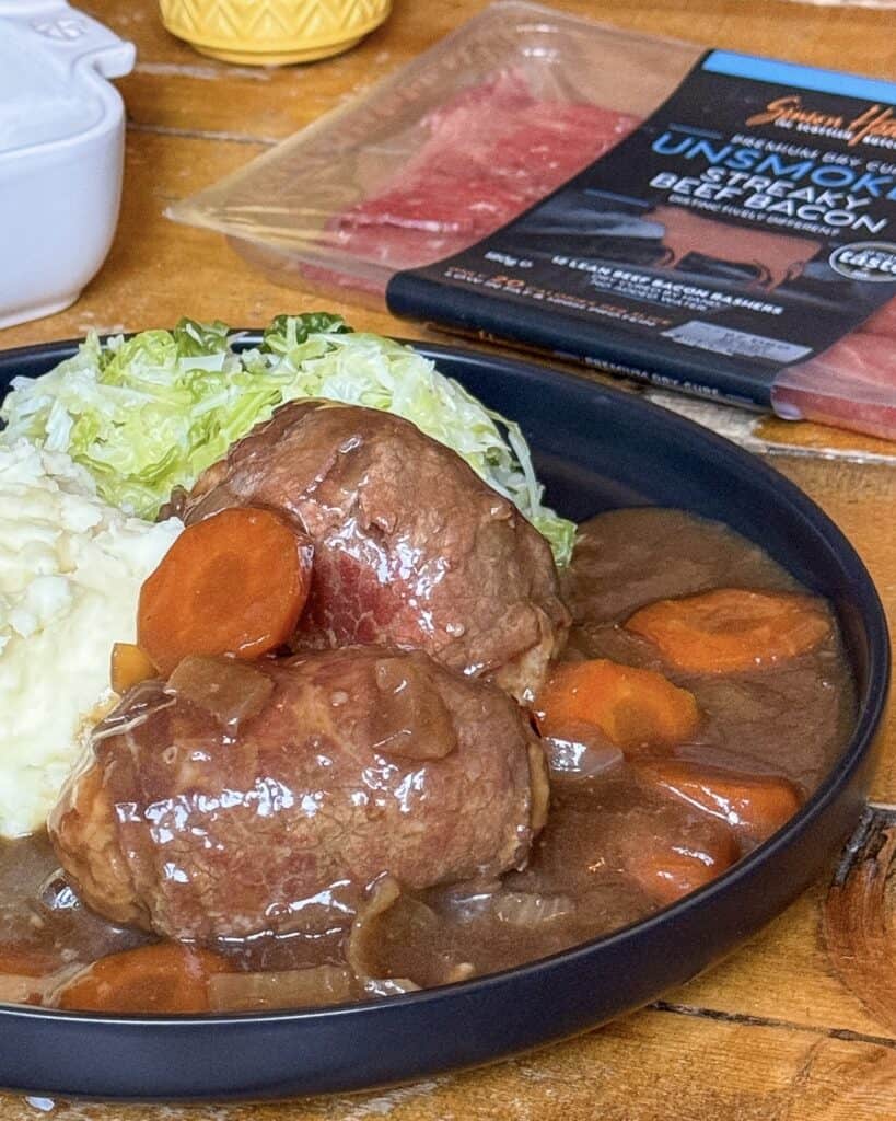 A plate with mashed potatoes, cabbage, and two beef olives in gravy with carrots. In the background, there is a package of uncooked steak and bacon slices on a wooden table.