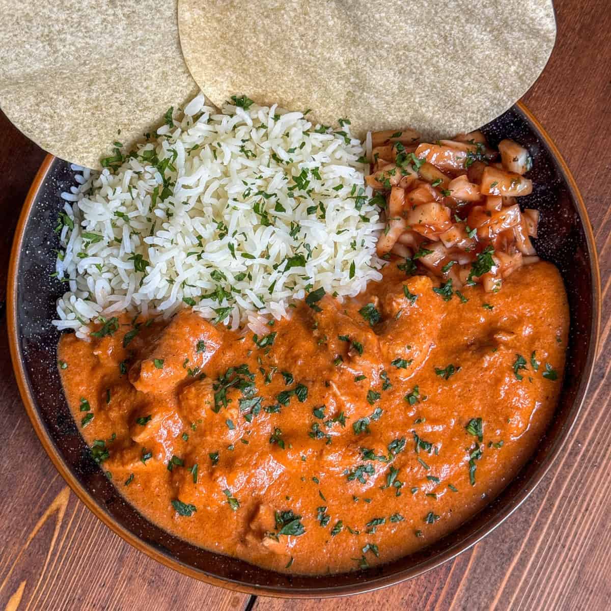 A plate with white rice, chicken makhani curry with herbs, spiced onions, and two poppadoms on the side, served on a dark dish atop a wooden table.