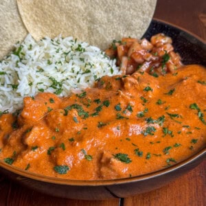 A bowl of creamy chicken makhani curry with chunks of meat, served with white rice, salsa, fresh herbs, and two corn tortillas on the side.