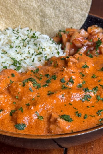 A bowl of creamy chicken makhani curry with chunks of meat, served with white rice, salsa, fresh herbs, and two corn tortillas on the side.
