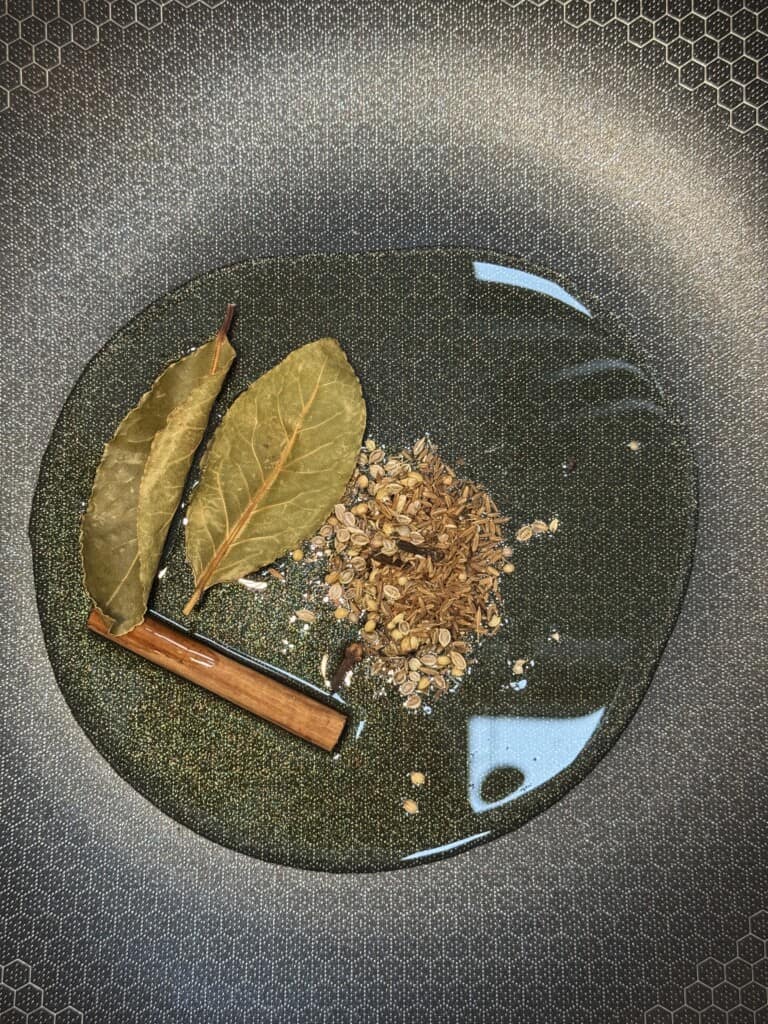 Whole spices including two bay leaves, a cinnamon stick, and a pile of cumin seeds are heating in oil inside a textured nonstick pan.
