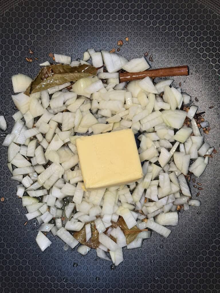 Finely chopped onions and a square of butter cooking in a pan with whole spices, including a cinnamon stick and bay leaves, on a textured, dark surface.