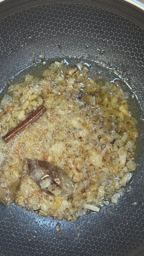 Chopped onions being sautéed in oil with whole spices, including a cinnamon stick and bay leaf, in a nonstick pan. The onions are turning golden brown.