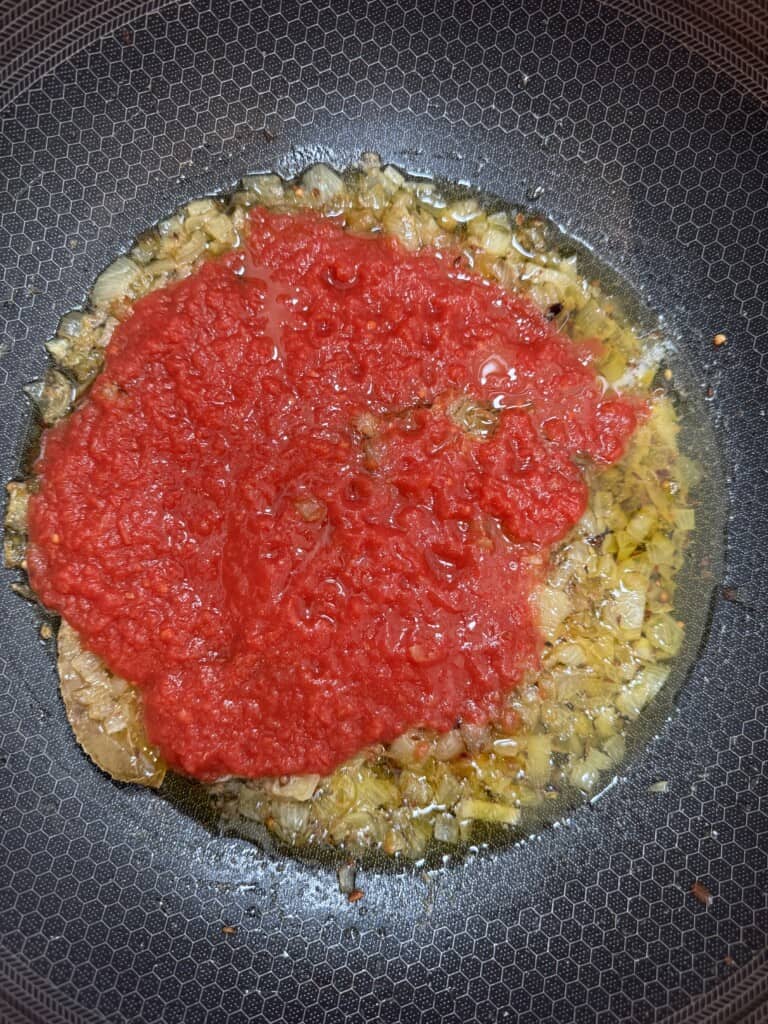 Chopped onions sautéing in oil in a pan, with a layer of pureed tomatoes added on top, ready to be cooked together.