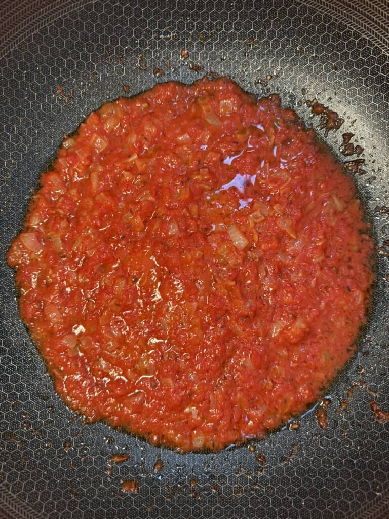 Chopped onions and blended tomatoes sautéing in oil in a black nonstick pan, forming a red sauce.