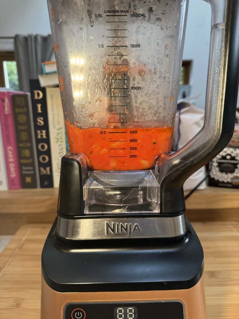 A close-up of a Ninja blender blending a red sauce, likely tomato-based. The blender sits on a wooden counter, with cookbooks, including Dishoom, visible in the background.