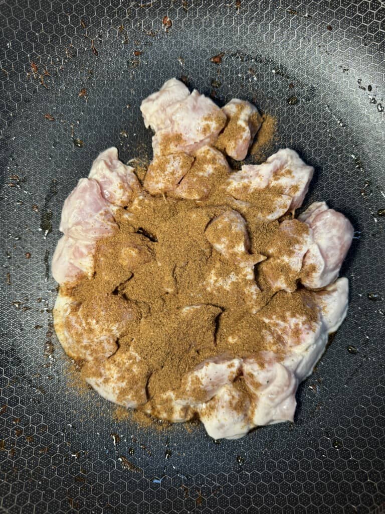 Raw, cubed chicken pieces topped with brown spice powder sit in the center of a textured, non-stick frying pan, ready to be cooked.