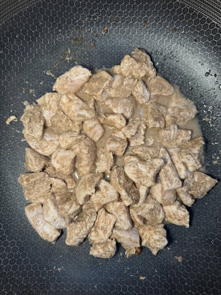 Small pieces of raw chicken breast cooking in a frying pan, lightly seasoned and beginning to brown, with some liquid visible in the pan.