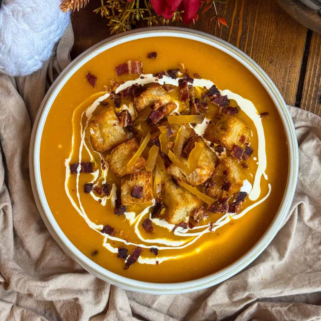 A bowl of creamy butternut squash and ginger soup topped with croutons, crispy bacon bits, a swirl of cream, and garnished with herbs, served on a wooden table with a beige cloth underneath.