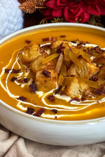 A bowl of butternut squash and ginger soup, garnished with croutons, crispy bacon bits, cheese shreds, and swirls of cream. The bowl sits on a beige cloth, with red flowers and a white knitted item in the background.