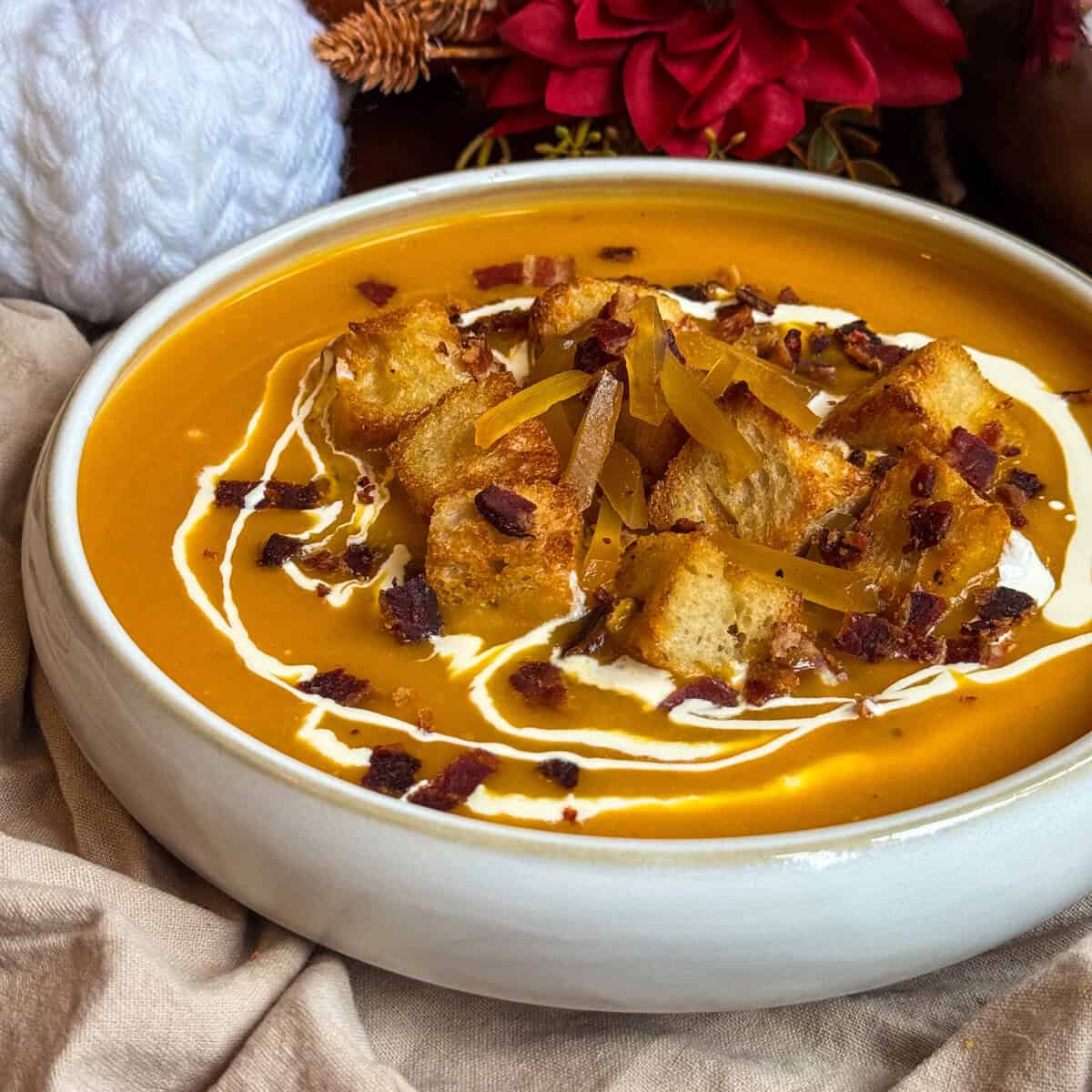 A bowl of butternut squash and ginger soup, garnished with croutons, crispy bacon bits, cheese shreds, and swirls of cream. The bowl sits on a beige cloth, with red flowers and a white knitted item in the background.