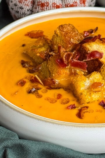 A close-up of creamy butternut squash coconut and chilli soup topped with golden croutons and crispy bacon bits, served in a white bowl on a dark green cloth.